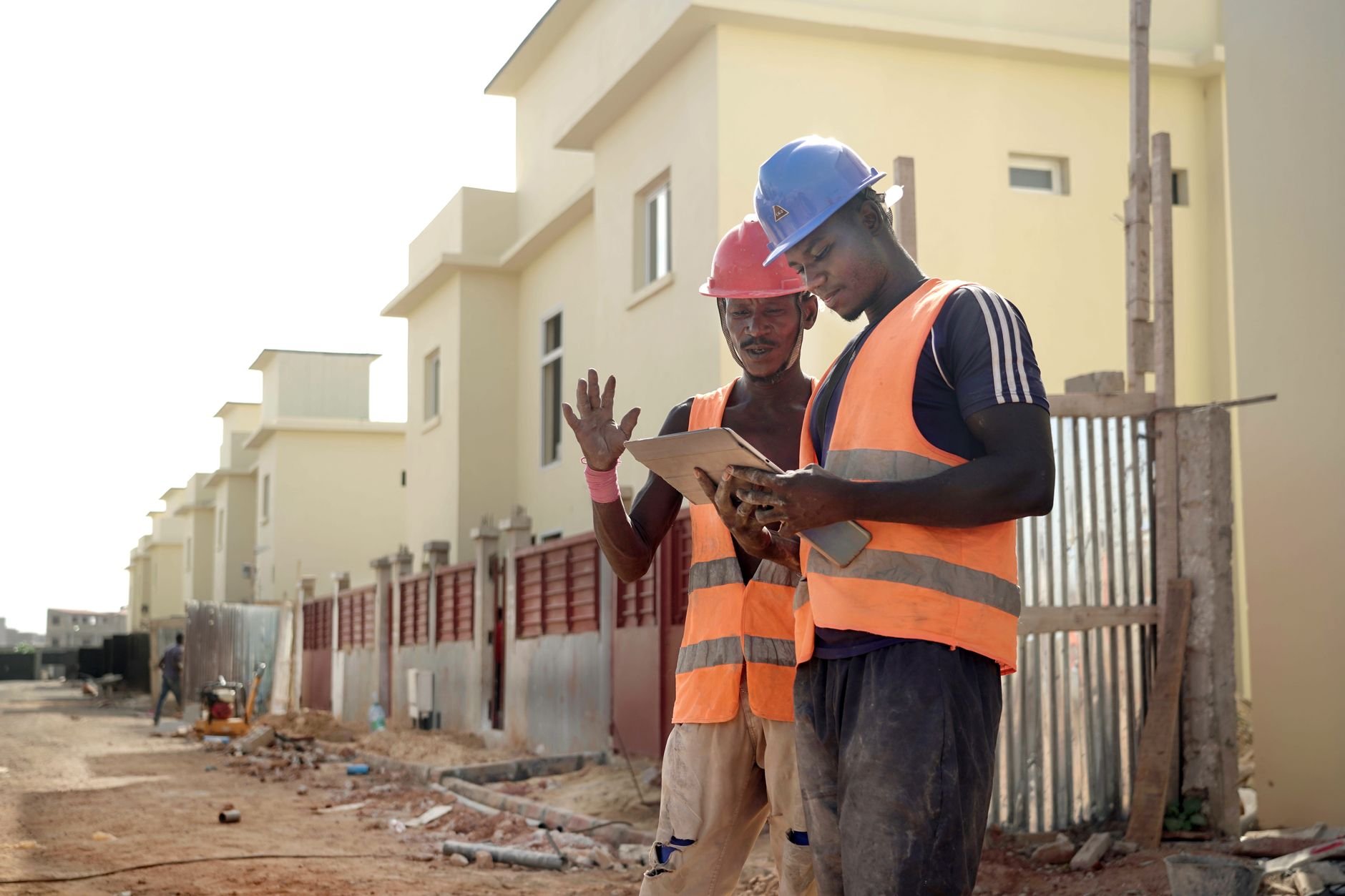 Two construction workers in helmets and vests review blueprints on a site.